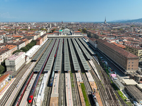Aerial view of Porta Nuova railway station with multiple platforms, red high-speed trains, and the city skyline under a clear blue sky Turin, Piedmont, Italy.