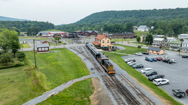 A train moves along the tracks in a small town. Buildings and parked cars are seen nearby, while green hills rise in the background. The scene captures daily life in this area.