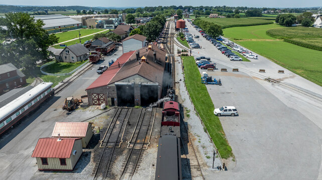 A steam train moves along the tracks at a station surrounded by buildings and vehicles. Nearby fields show green crops and open space under a clear sky. People are visible near the station.