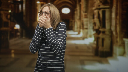 Fototapeta premium Woman covers mouth with hands and wears glasses in ornate building corridor with arches and columns; illness concern.