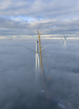 Aerial view of wind turbines and a yellow lattice boom crane emerging from a thick blanket of white fog under a cloudy sky.