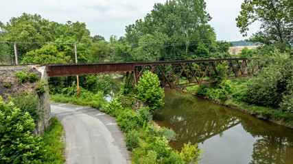 Fototapeta premium A metal railroad bridge spans over a small river next to a winding road. Lush green trees line the banks of the river. The sun shines down on the scene, showing a peaceful afternoon.