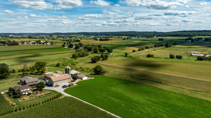 A wide view of a rural area with green fields, farm buildings, and a clear sky. The setting is peaceful with a mix of crops and open land under clouds in the sky. © Greg Kelton