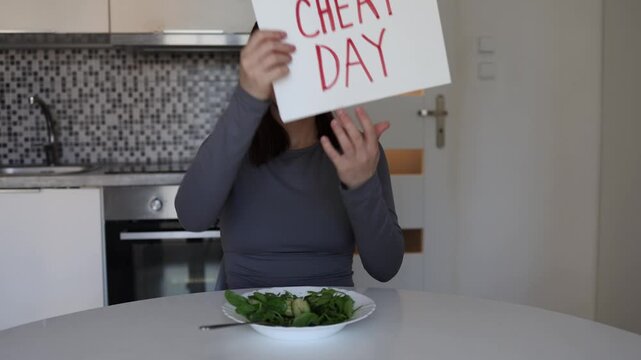 Young woman breaks healthy diet routine enjoying chocolate during cheat day while holding motivational sign and laughing alone in modern kitchen at home