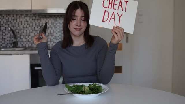 Young woman declares cheat day with no food restrictions ignoring healthy salad and choosing favorite chocolate snack while relaxing alone in cozy home kitchen
