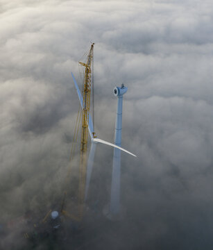 Aerial view of a wind turbine under construction with a tall yellow crane rising above a thick layer of white fog in Netherlands.