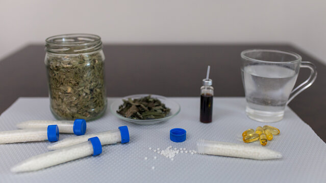 Close-up of homeopathic and natural medicine items: glass jar of dried herbs, tubes with pellets, yellow capsules and glass of water. Holistic remedies and alternative healthcare.