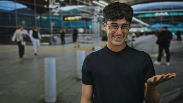 Man gesturing palm up at airport terminal with outstretched hand and eyeglasses, smiling in a casual pose; curiosity travel welcome.