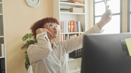 Woman holding smartphone smiling and raising arm to take selfie at desk with computer and bookshelves in building  workday joy. © Krakenimages.com