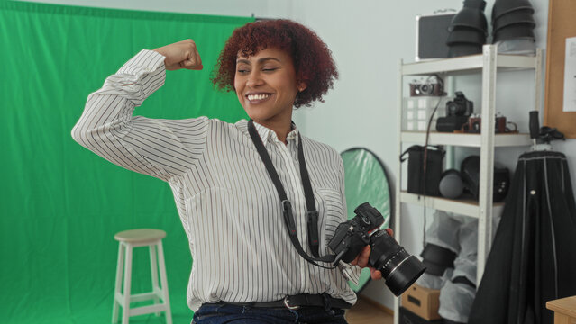 Woman photographer holding camera and strap flexes arm showing bicep in studio with green backdrop, stool, shelving, lights and reflector; confidence.