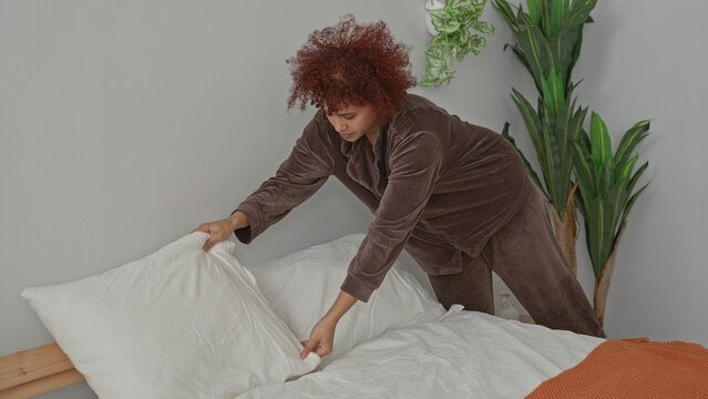 Woman places pillow on bed and smooths white sheets while tucking an orange throw beside a wooden headboard in a building bedroom; calm routine.