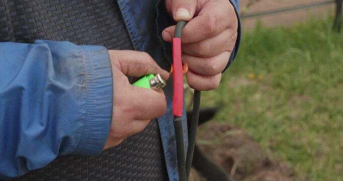 Worker's hands using a lighter to heat and shrink a red insulating tube over a repaired electrical cable, restoring the connection and providing a secure, waterproof seal for safety.