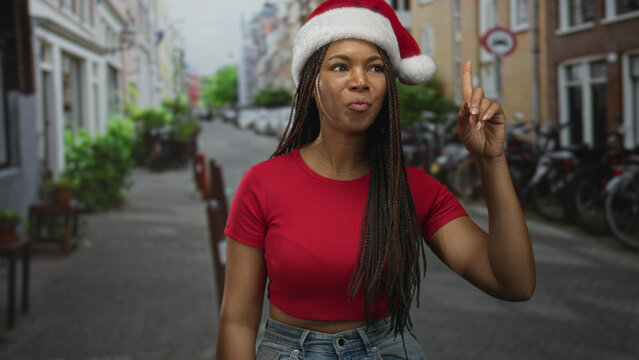 Woman in santa hat and red cropped shirt pointing finger upward on a narrow cobbled street lined with bicycles and brick buildings, wearing jeans and long braids; playful holiday idea.