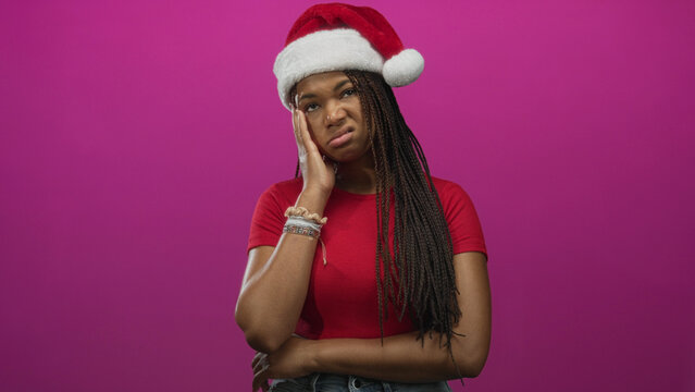 Woman with santa hat touching face with hand and grimace wearing cropped red top in pink studio; annoyance holiday.