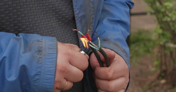 Close-up of a worker's hands using a lighter to heat shrink an insulating tube on a repaired electrical wire, protecting the connection of the copper cables outdoors.