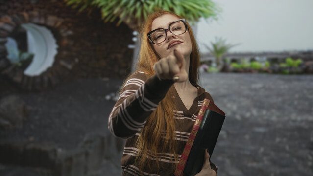 Young redhead woman points index finger at camera while clutching a hardcover book in front of an urban building rooftop; confidence attitude.