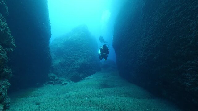  scuba divers exploring around a reef underwater deep blue water big rocks and bubbles ocean scenery 