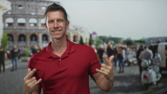 Man holds up two fingers in front of the coliseum building showing forearms and a red polo, smiling at camera; adventure playful.
