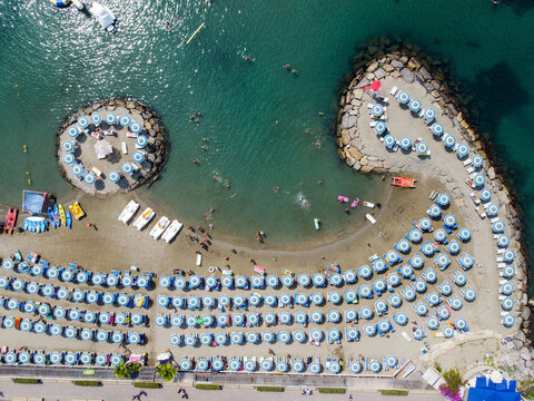 Aerial view of rows of blue and white umbrellas on a sandy beach with people swimming and relaxing near stone breakwaters San Lorenzo, Liguria, Italy.