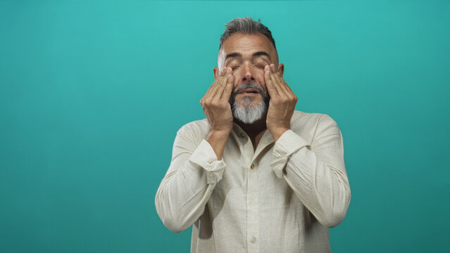 Man middle aged with gray beard and beige shirt covering his eyes with hands in studio against teal backdrop; surprise.