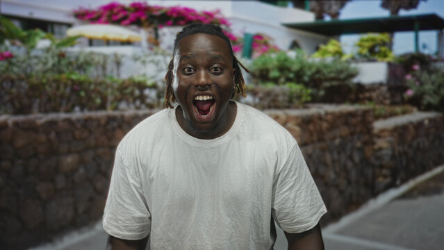 Man smiling showing teeth on street with stone wall and flowering terrace, wearing white tshirt; joyful optimism.
