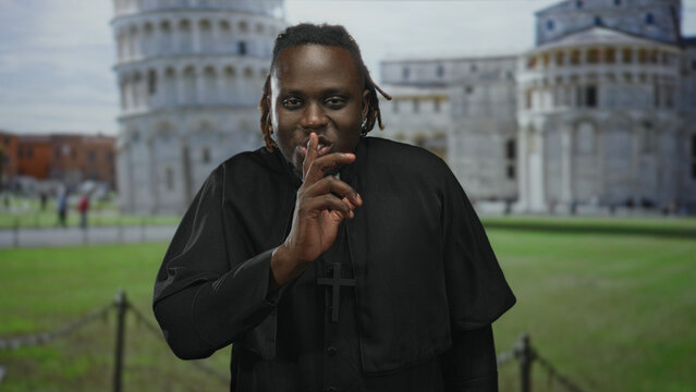 Man in clerical robes wearing a prominent cross necklace, finger to lips for silence in building at the leaning tower of pisa plaza; silence reflection.