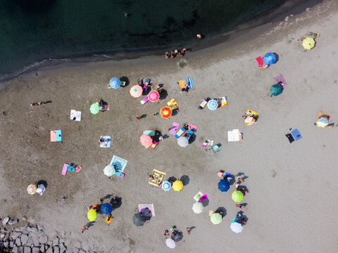 Aerial view of colorful beach umbrellas and towels scattered on the sand with people sunbathing near the dark sea water in San Lorenzo, Liguria, Italy.