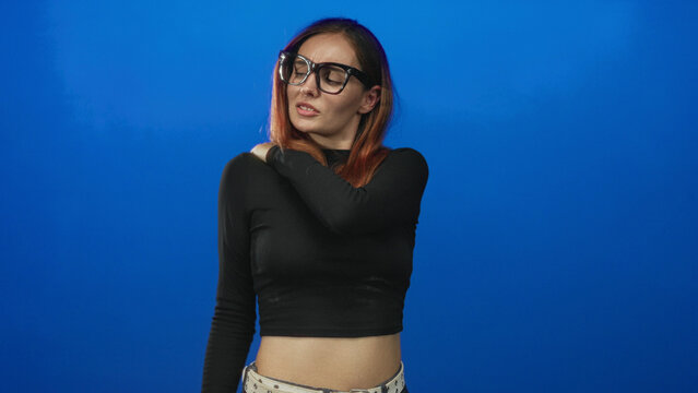 Young redhead woman wearing glasses and black cropped top holds her shoulder with a pained grimace in a studio; discomfort.