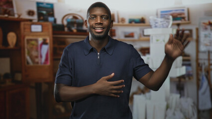 Fototapeta premium Man raises right hand with open palm and holds left hand to chest while standing in an art studio with shelves and display boxes; confidence friendliness.