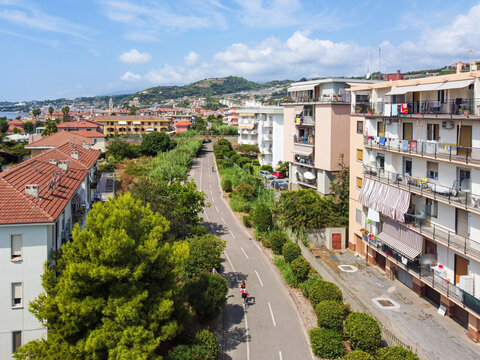 Aerial view of a paved cycle path lined with green trees and multi-story residential buildings under a bright blue sky with distant hills in San Lorenzo, Liguria, Italy.