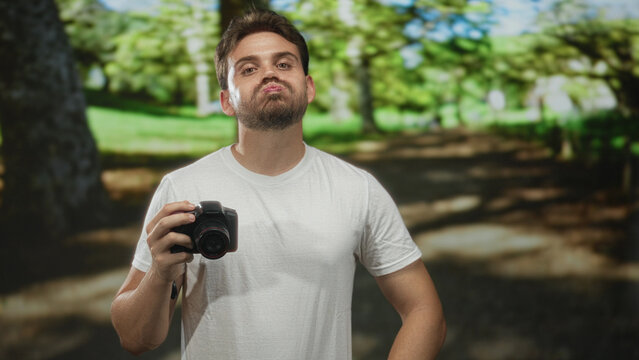 Man holding camera with one hand and smirking in forest park setting wearing white tshirt and beard visible; playful curiosity.