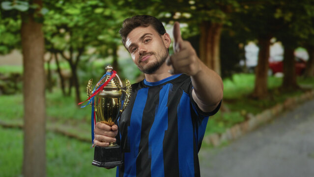 Man holding trophy points finger in forest wearing blue striped jersey in green park setting; pride victory.