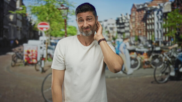 Man touching neck with hand and wry half smile beside bicycles by a canal on a street in amsterdam; awkwardness uncertainty.