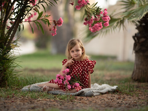 Beautiful child sitting on blanket under blooming pink oleander bush in park warm summer day