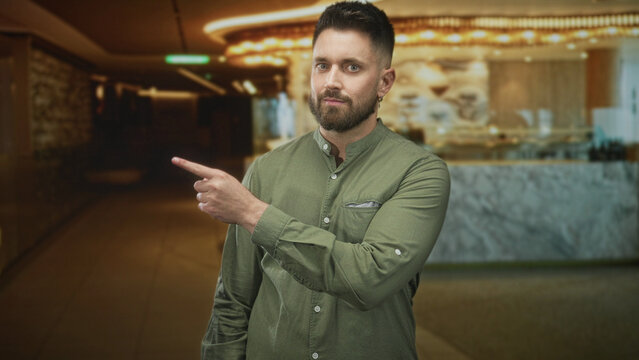 Man with beard pointing index finger toward corridor in building lobby beside marble counter wearing green shirt and earring; confidence.