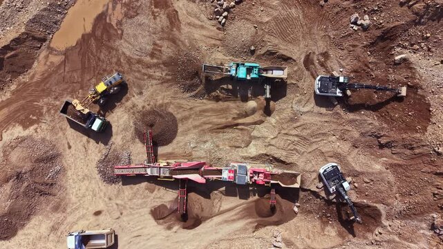 Workers operate heavy machines to mine and move earth and rocks on a construction site. The scene shows several types of machinery at work on the ground.