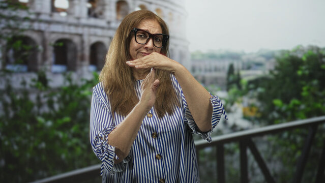 Woman with glasses makes timeout gesture with hands on a balcony overlooking ancient building colosseum; travel memory playful.