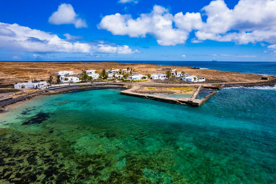 The scene shows aerial view of La Graciosa Canary island near Lanzarote with clear turquoise water, small village, rocky coastline, and white buildings under a blue sky with clouds