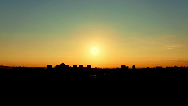 The sun rises above the city skyline, casting light on the buildings. The bright sky changes colors as the day begins. The scene shows a quiet morning with no movement.