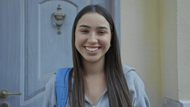 Young hispanic woman student wearing gray hoodie with blue backpack smiles at camera showing teeth and dimples at a building entrance doorway; optimism new start.
