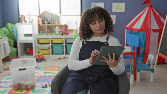 Woman teacher tapping tablet while seated in a colorful school classroom with toys and play tent; focus lesson planning.