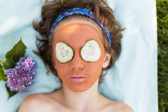 Overhead view of a young boy lying on a towel in a garden with a face mask on and cucumber slices on his eyes