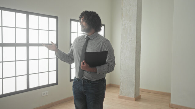 Man holding black folder gestures with open hand and points with other hand in empty building interior, smiling and presenting the space; confidence trust.