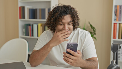 Man holding smartphone with thumb tapping screen while smiling at notifications, seated by bookshelves in a building  joy connection. © Krakenimages.com