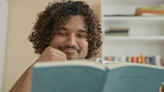 Young indian man reading teal book, hand on chin while turning page in a building with bookshelf and wall clock visible; contemplation.