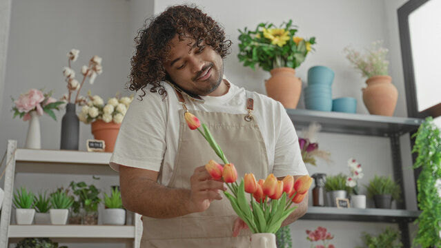 Man arranging orange tulips with phone held at shoulder while adjusting stems in apron inside a flower shop building; calm focus.