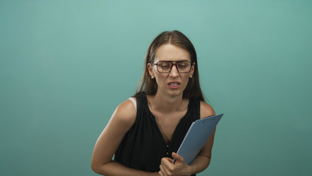 Woman wearing glasses holding a blue clipboard and grimacing while clutching it to her chest in teal studio; disgust.