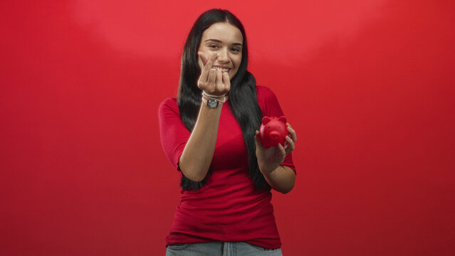 Young woman holding red piggybank and making finger heart gesture in a studio with bright red backdrop; financial optimism.