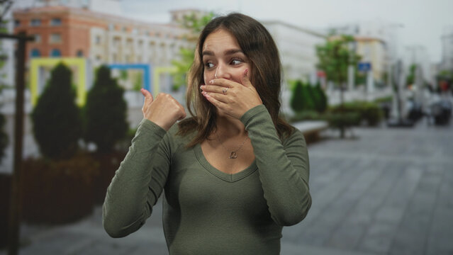 Young hispanic woman wearing green top and heart necklace, covering mouth with hand and pointing thumb on street; surprise.