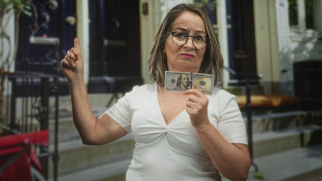 Woman grey hair holds a hundred dollar banknote and points finger upward at a building stoop with raised arm; financial frustration concern.